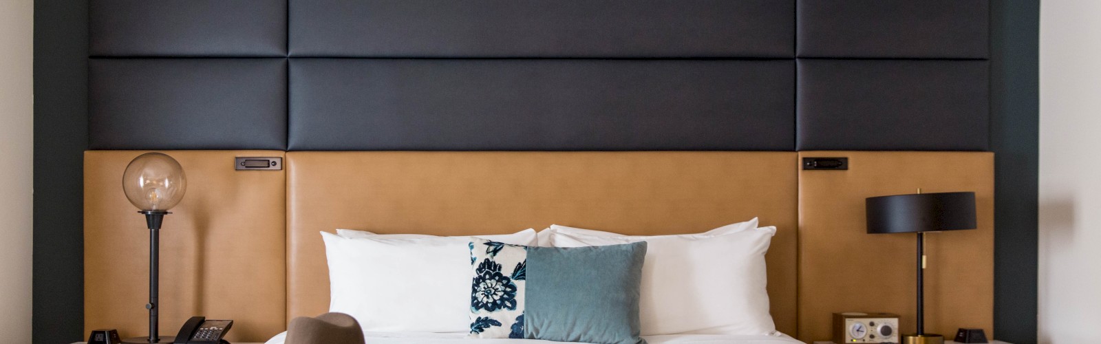 A neatly made bed with pillows, a hat on the blanket, and two bedside tables with lamps and other items in a modern bedroom.