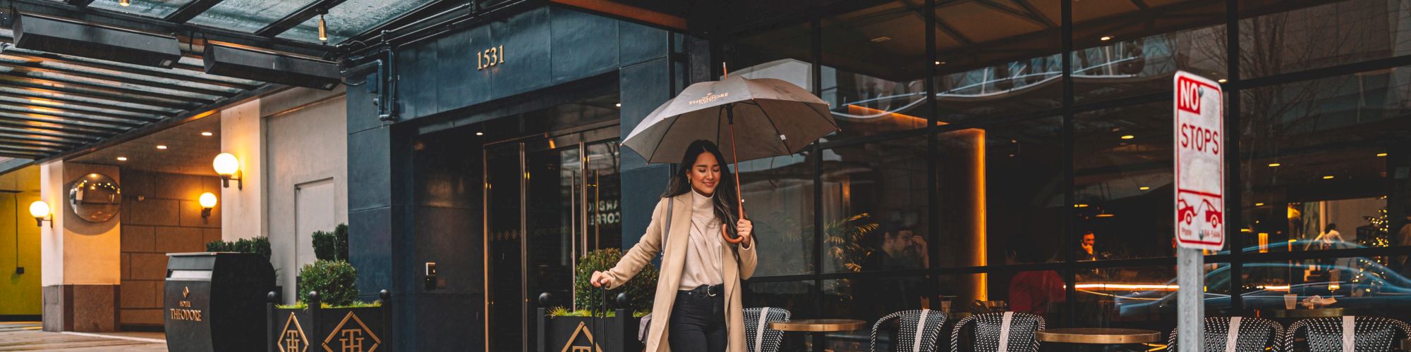 A woman holding an umbrella is walking with rolling luggage outside Hotel Theodore on a rainy day. She is dressed in a long coat and casual attire.