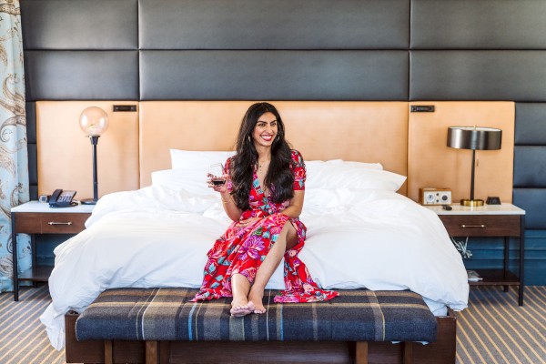 A woman in a floral dress is sitting on a bed holding a glass, in a modern hotel room setting.