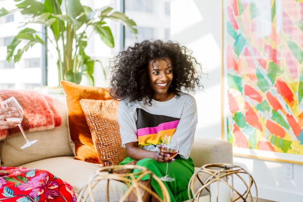 A person smiling on a couch, holding a glass, with colorful pillows and artwork in the background. Bright and cheerful atmosphere.