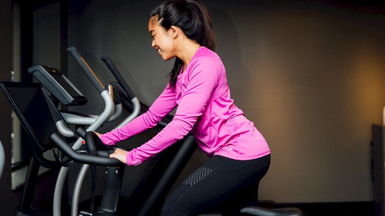 A woman is riding a stationary bike in a gym, wearing a pink long-sleeve shirt and black pants, with other exercise equipment in the background.