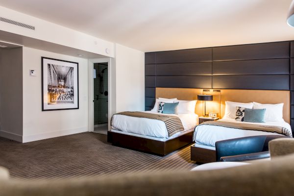 The image shows a modern hotel room with two double beds, a nightstand with a lamp, a framed photograph, and a chair. The decor is contemporary.