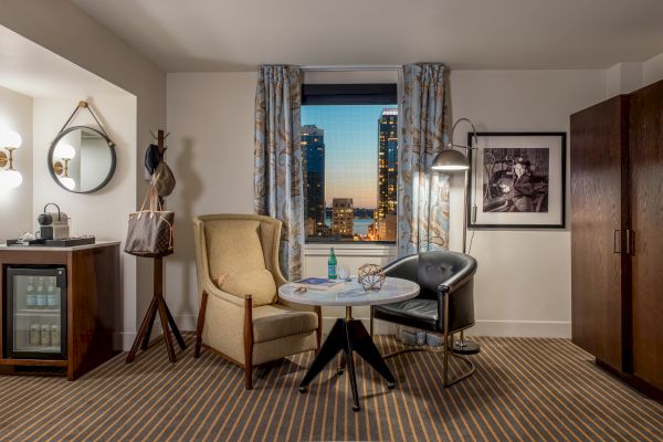 A cozy hotel room featuring a small table, two chairs, a mini-fridge, wardrobe, wall art, and a window with a cityscape view at dusk.