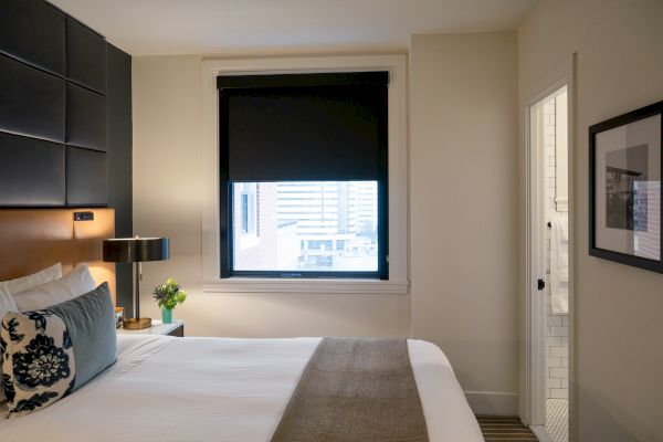 A modern hotel room features a neatly made bed, a window with a black roller shade, a nightstand with a lamp and plant, and a doorway to a bathroom.