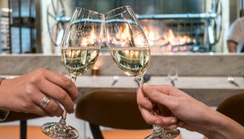 Two hands clink wine glasses in a cozy restaurant setting with a warm fireplace in the background, creating a celebratory mood.
