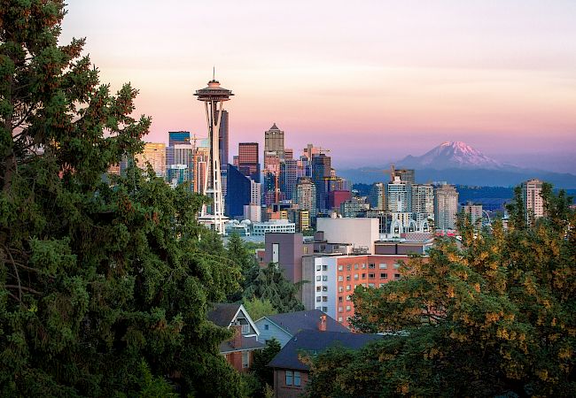 The image shows the Seattle skyline with the Space Needle, skyscrapers, and Mount Rainier in the background, framed by trees in the foreground.