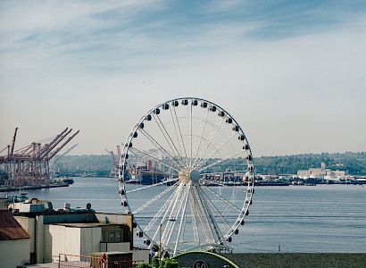 The image shows a Ferris wheel near a waterfront with cranes and buildings in the background under a partly cloudy sky.