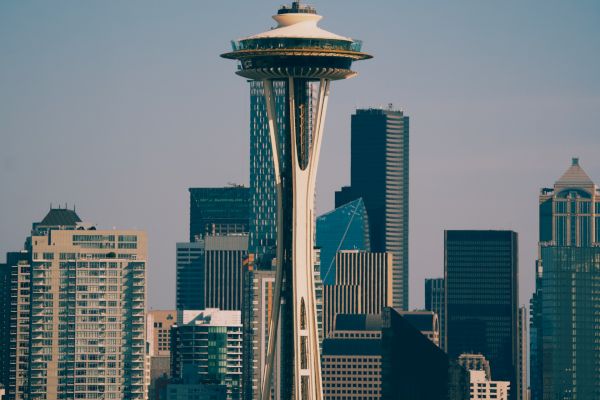 The image shows the Space Needle with the Seattle skyline in the background on a clear day. The skyscrapers are prominent against the blue sky.