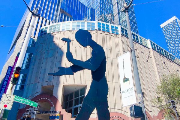 The image shows a large silhouette sculpture of a man with a hammer in front of a modern building under a clear blue sky, with people walking by.