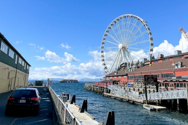 A coastal scene with a ferris wheel, buildings, parked cars, and a railing along the water under a clear blue sky.
