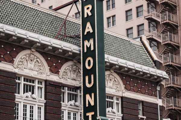 The image shows the front of the Paramount Theater with a large vertical sign displaying its name and a marquee listing upcoming shows.