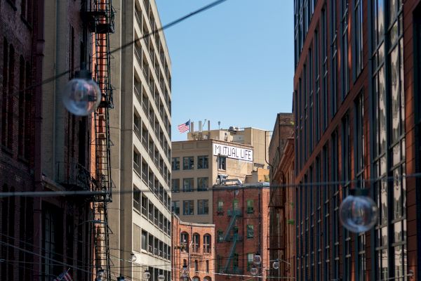 This image shows an urban alleyway with hanging light bulbs, surrounded by tall buildings, and a clear blue sky in the background.