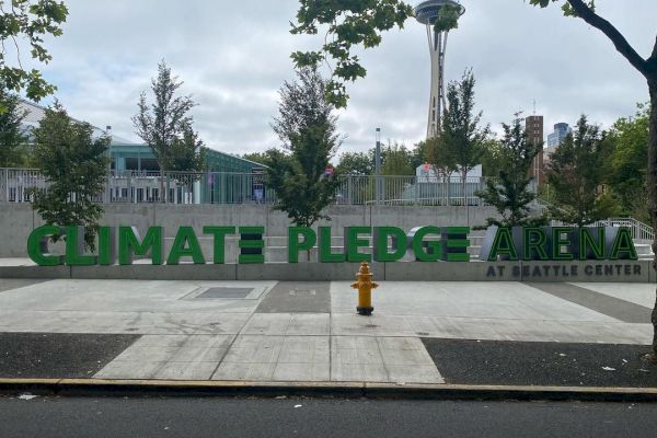 The image features the Climate Pledge Arena in Seattle. The Space Needle is visible in the background, with surrounding trees and a fire hydrant.