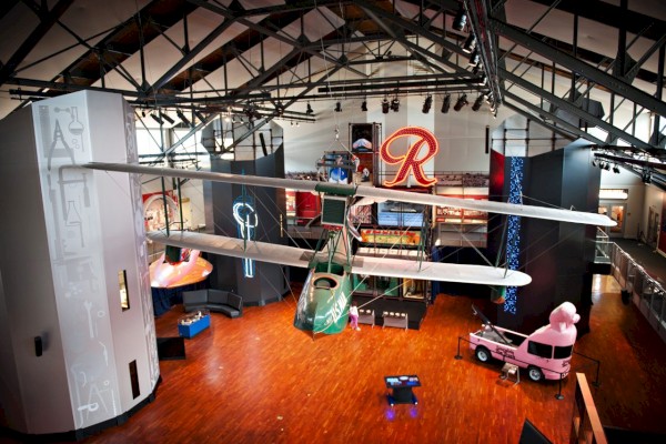 The image shows a well-lit museum exhibit hall featuring a suspended green airplane, various displays, a large 