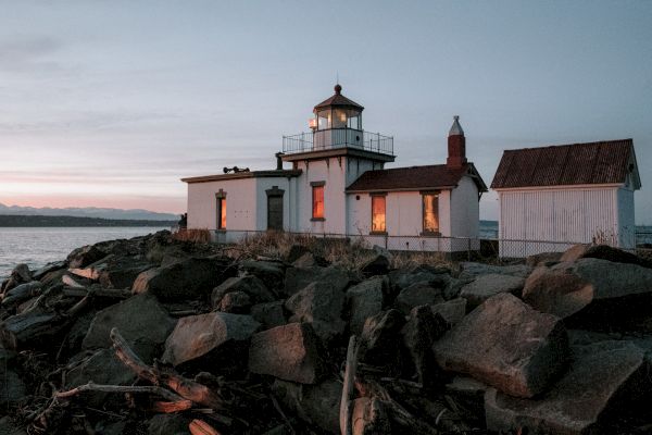A lighthouse with adjoining buildings stands on a rocky shore during dusk, with warm lights glowing from the windows, and a calm sea in the background.