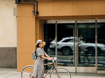 A person wearing a hat and checkered dress stands beside a bicycle in front of Hotel Theodore's entrance, with cars reflected in the glass doors.