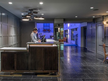 A modern hotel lobby with a person at the front desk, dim lighting, paneled walls, and a corridor leading to other areas of the building.