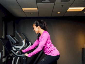 A person in a pink top is riding an exercise bike in a gym. The background includes other gym equipment and a wall-mounted television.