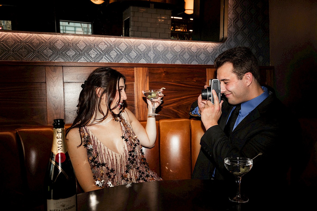 A woman toasts with a drink while a man photographs her in a bar. A bottle and a glass are on the table.