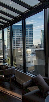 A modern indoor lounge area with large windows offers a panoramic view of city skyscrapers and buildings, with seating and tables along the side.