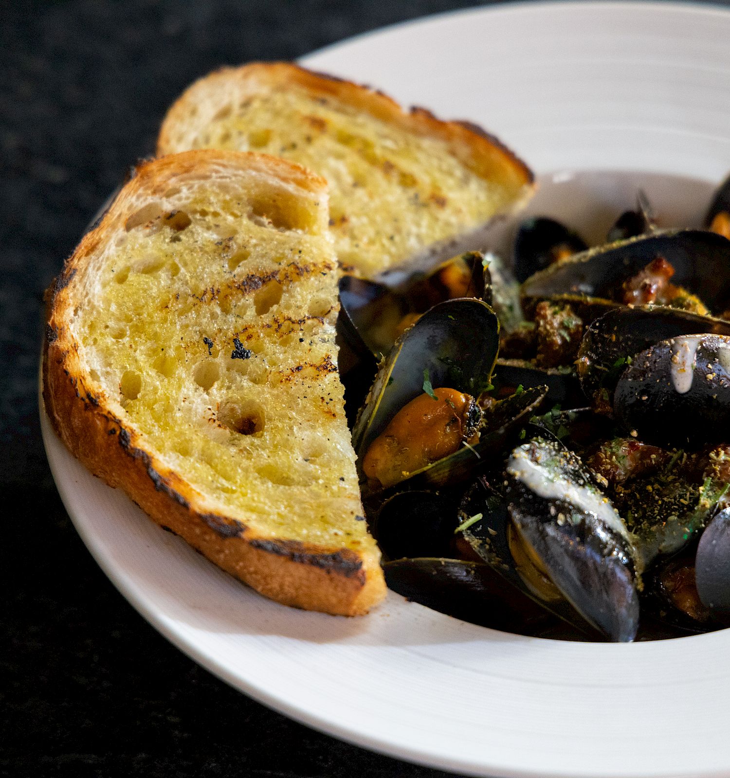A white plate with cooked mussels and two slices of toasted bread on a dark surface.
