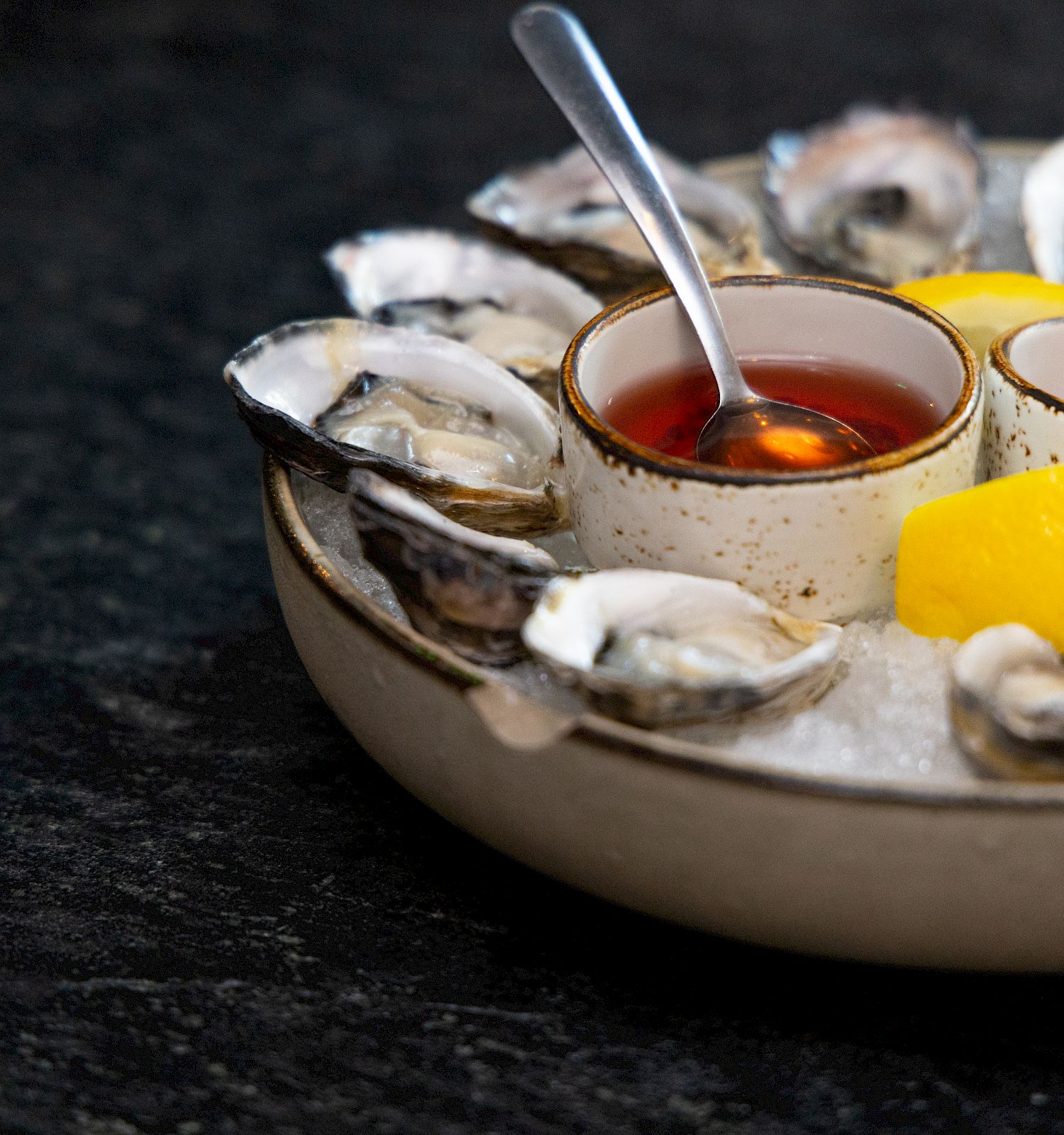 The image shows a dish of oysters on ice, accompanied by lemon wedges and two sauces in small bowls with spoons.