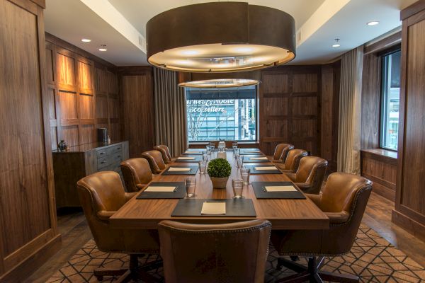 A well-furnished conference room with a long wooden table, leather chairs, a center plant, and overhead lighting, featuring large windows and wooden paneling.