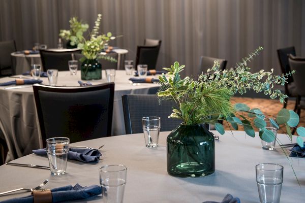 The image shows a dining setup with round tables draped in white tablecloths, green glass vases with greenery, and blue napkins, in an elegant setting.
