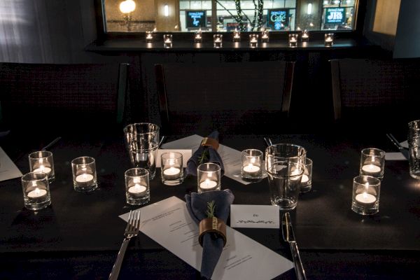 A dimly lit dining setup with numerous candles, glasses, napkins, cutlery, and menus arranged on a dark table in a cozy restaurant setting.
