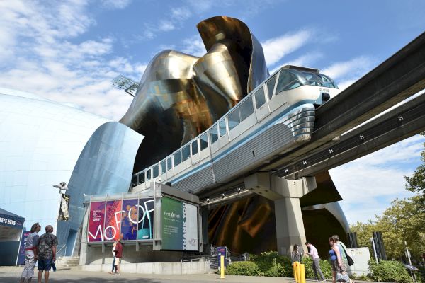 A futuristic monorail passes by a modern, abstract building with a colorful sign and people walking around, set against a bright, partly cloudy sky.