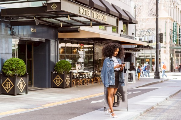 A woman stands outside Hotel Theodore reading a menu board, with nearby greenery and pedestrian activity in the background.