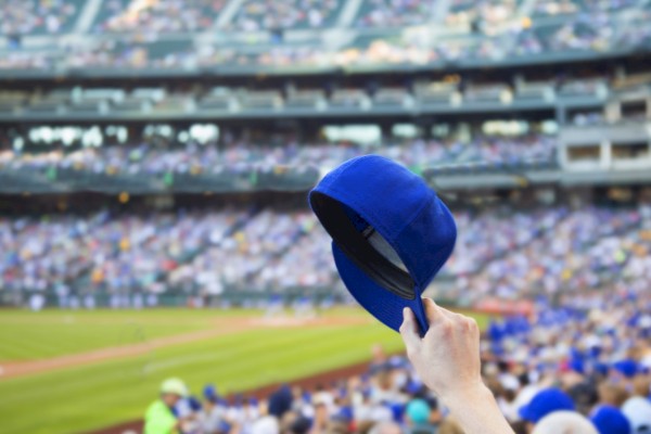 A person holds up a blue baseball cap in a stadium full of spectators. The crowd and baseball field are visible in the background.