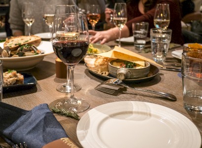 A table setting with food, drinks, wine glasses, and people dining, with cutlery, plates, and a napkin visible in a cozy restaurant atmosphere.