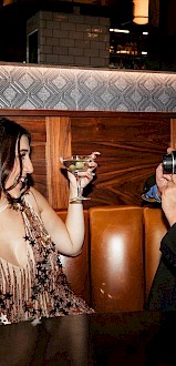 A woman holds a drink while a man photographs her in a cozy bar setting. A champagne bottle is on the table.