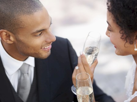 A couple dressed formally is smiling and raising champagne glasses, likely in a celebratory setting.