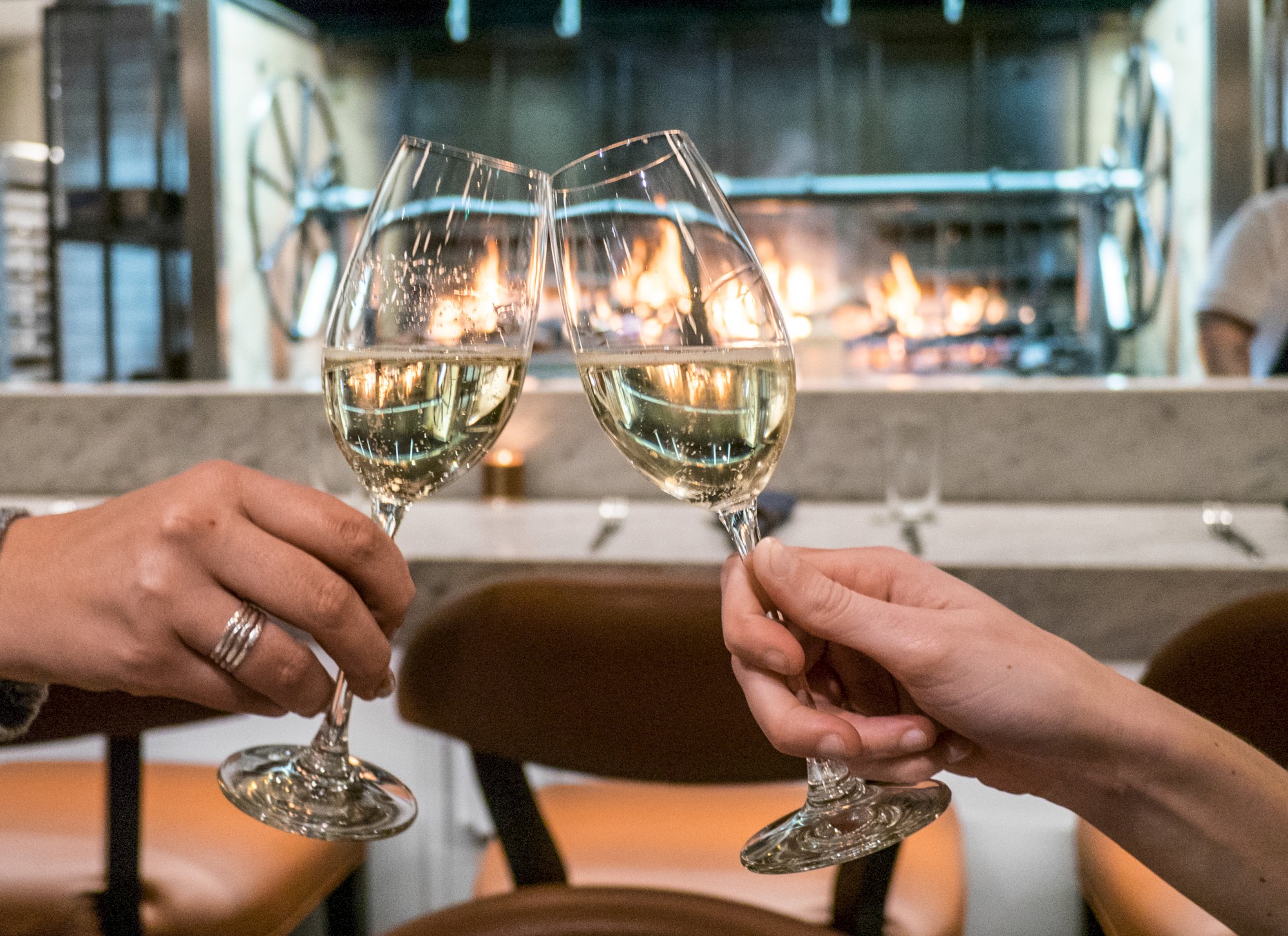 Two people clink wine glasses in a celebratory toast in front of a lit fireplace at a restaurant, with leather chairs and a cozy ambiance.