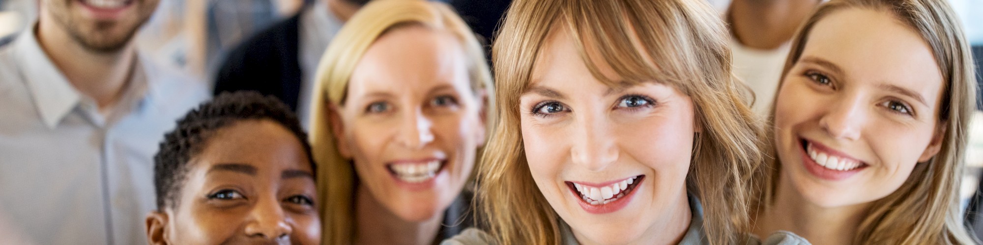 A group of eight people are taking a cheerful selfie, smiling and standing close together in what appears to be an indoor setting.