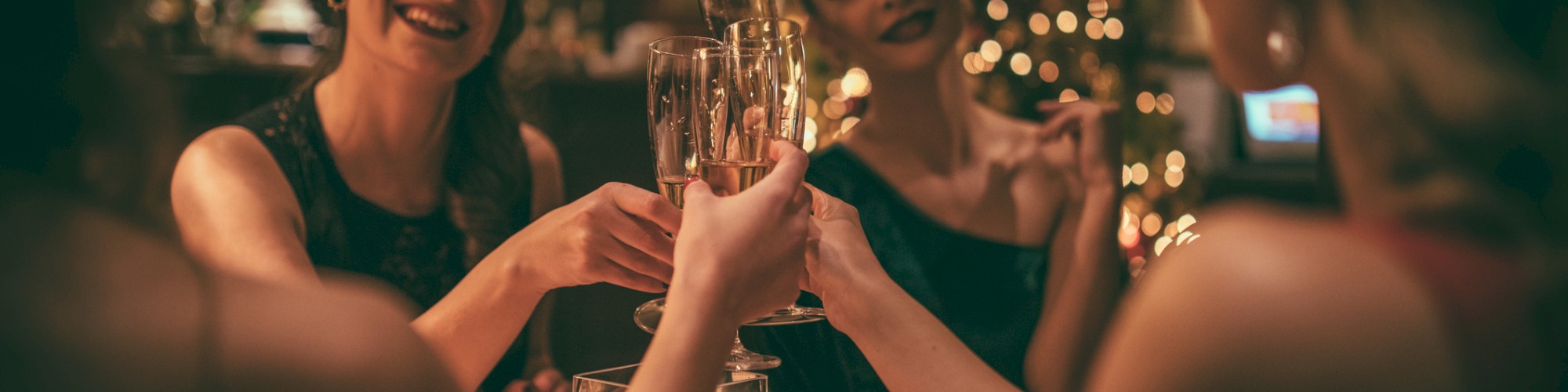 A group of women are raising glasses in a toast, sitting at a table in a warmly lit, festively decorated setting.