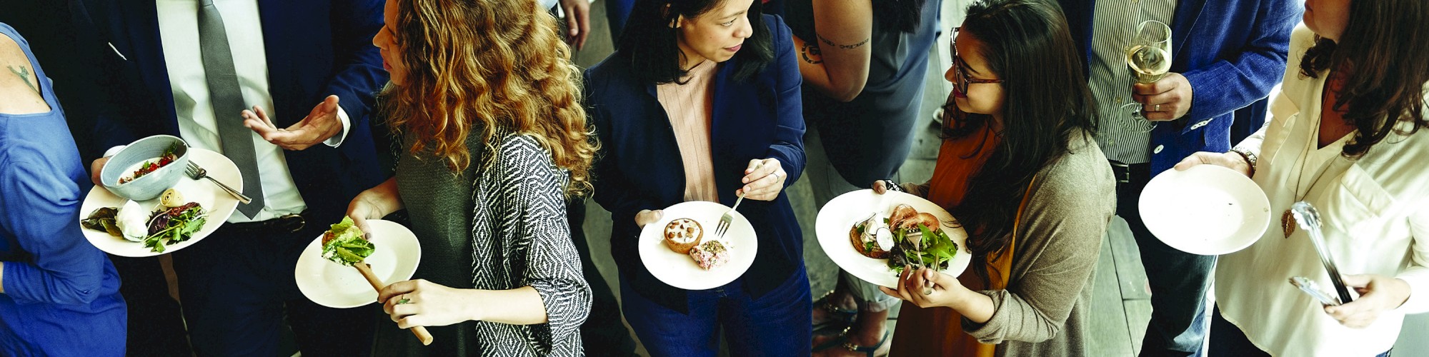 A group of people at a social gathering, standing around a table filled with various foods and drinks, chatting and enjoying the meal.