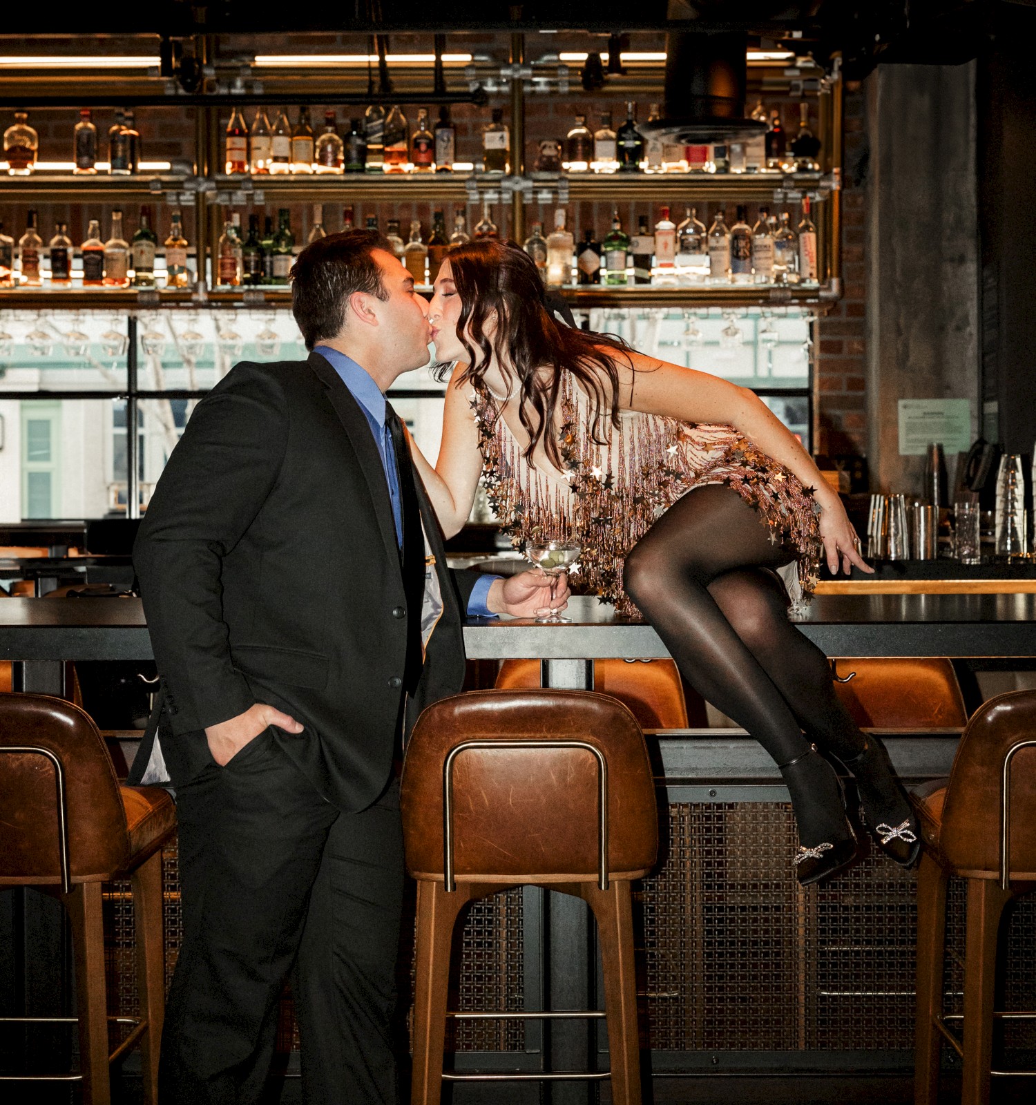 A man in a suit and a woman in a sparkly dress touching noses at a bar counter with shelves of bottles in the background.