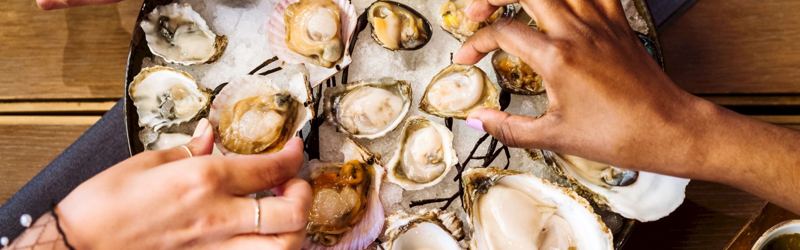 The image shows several hands reaching for a variety of oysters, served on a large tray with grilled lemon halves, on a table with various dipping sauces.