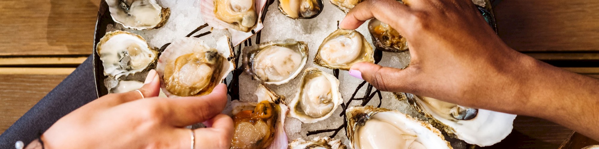 The image shows several hands reaching for a variety of oysters, served on a large tray with grilled lemon halves, on a table with various dipping sauces.