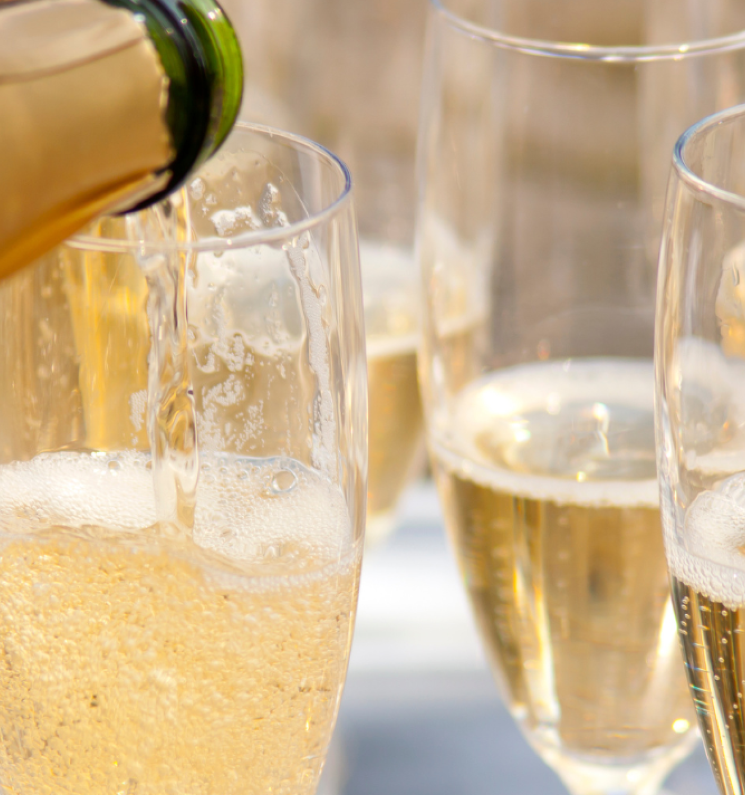 The image shows champagne being poured into several clear flute glasses, with bubbles forming as the liquid fills the glasses.