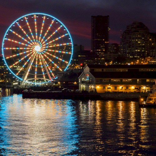 The image shows a lit-up Ferris wheel near a waterfront at dusk or night, with city buildings illuminated in the background.