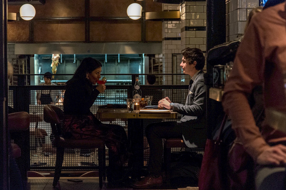 A man and woman dine at a restaurant table, chatting and enjoying drinks, with an open kitchen visible in the background.