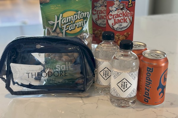 A clear Hotel Theodore bag, Hampton Farms peanuts, Cracker Jack boxes, bottled water, and Bodhizafa beer cans on a counter.